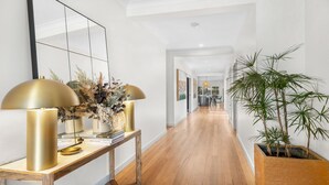 A hallway with a wooden floor, a console table with two gold lamps, and a large mirror. There are framed pictures on the wall and a potted plant on the right. - HUNTER DREAMING - THE VINTAGE HOLIDAY HOUSES (Pokolbin)