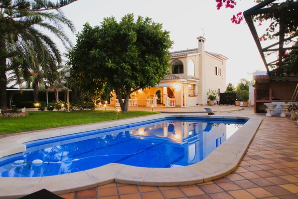 A swimming pool with a tiled deck, surrounded by a well-maintained lawn and a large tree.
