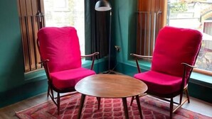 Ein Zimmer mit zwei roten Stühlen um einen Holztisch, einem gemusterten Teppich und Blick durch ein Fenster mit Vorhängen.
