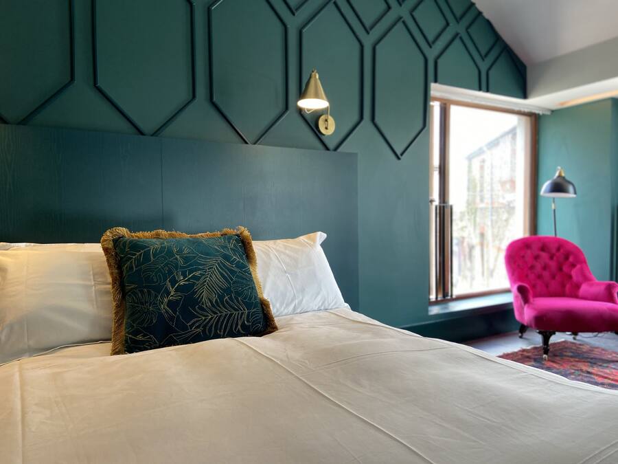Un dormitorio con cama, pared verde con diseños geométricos, ventana y sillón rosa.