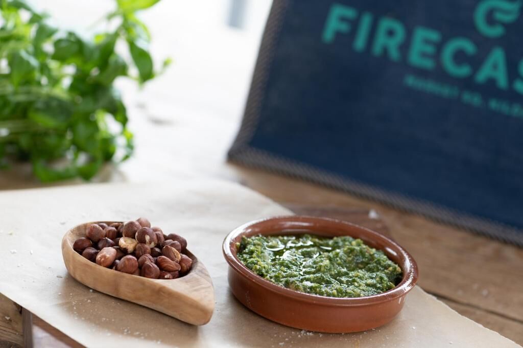 Un cuenco de madera con avellanas y un bol de terracota con pesto verde sobre una superficie de madera.