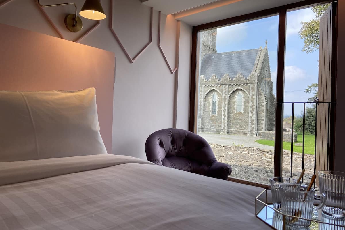 A bedroom with a large window, a bed, a chair, and a tray with glasses and a cup.