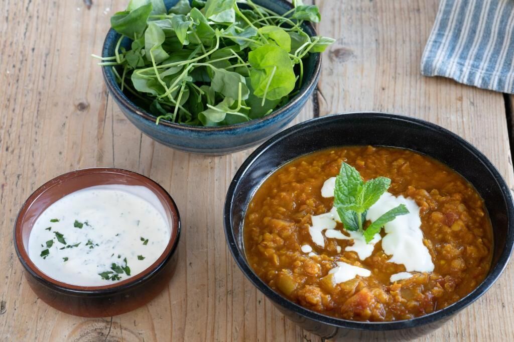 A bowl of lentil soup garnished with sour cream and mint, a bowl of yogurt with herbs, and a bowl of fresh greens on a wooden table.