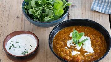 Un tazón de sopa de lentejas adornado con crema agria y menta, un tazón de yogur con hierbas y un tazón de verduras frescas sobre una mesa de madera.