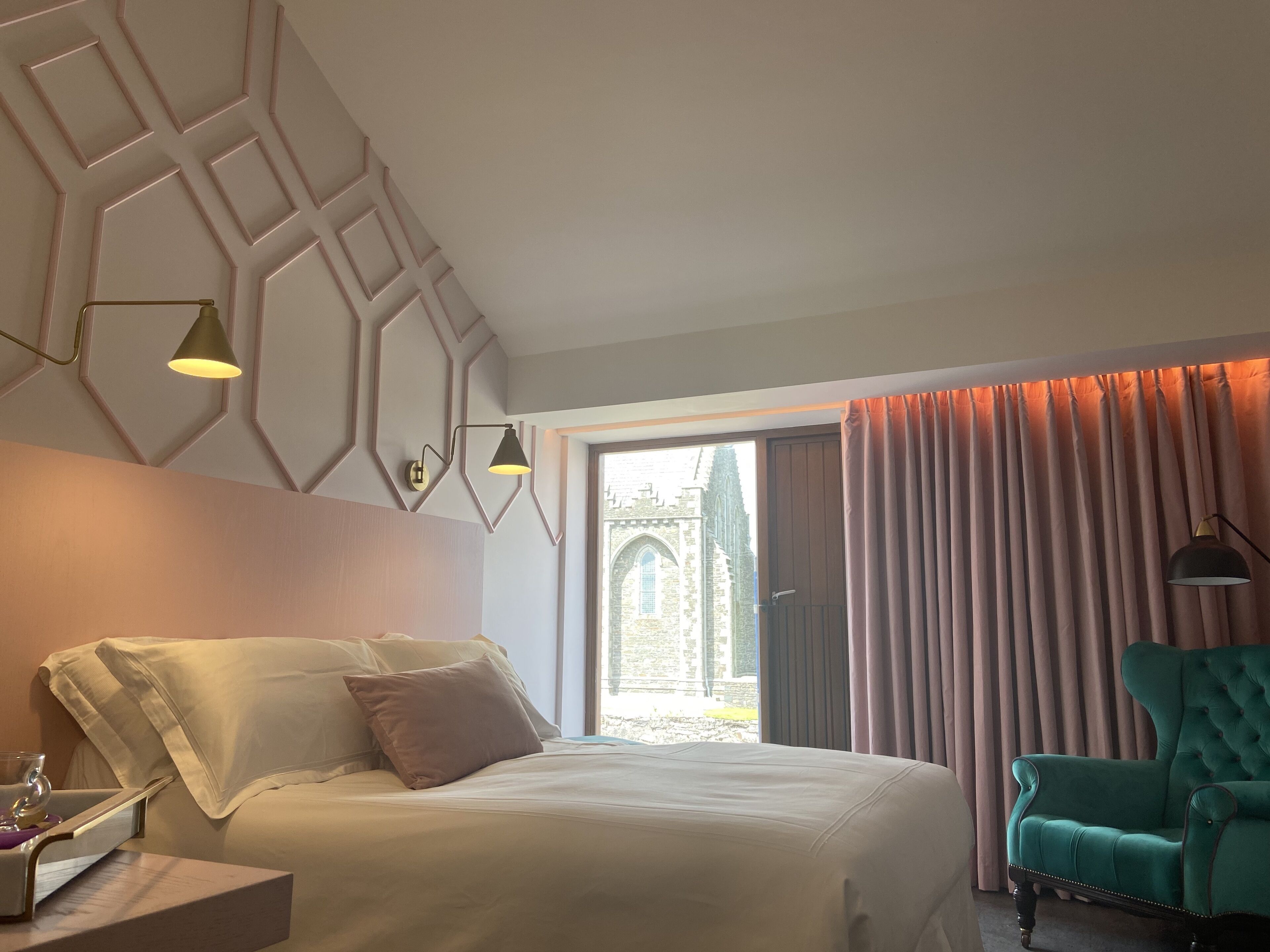 A hotel room with a bed, a nightstand, a green armchair, and a window with curtains.