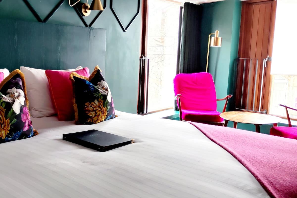 A hotel room with a large bed, multiple pillows, a small table, a chair, and a view of the outdoors.