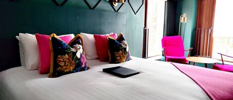 Habitación de hotel con una cama grande, varios almohadones, una mesita pequeña, una silla y vistas al exterior.