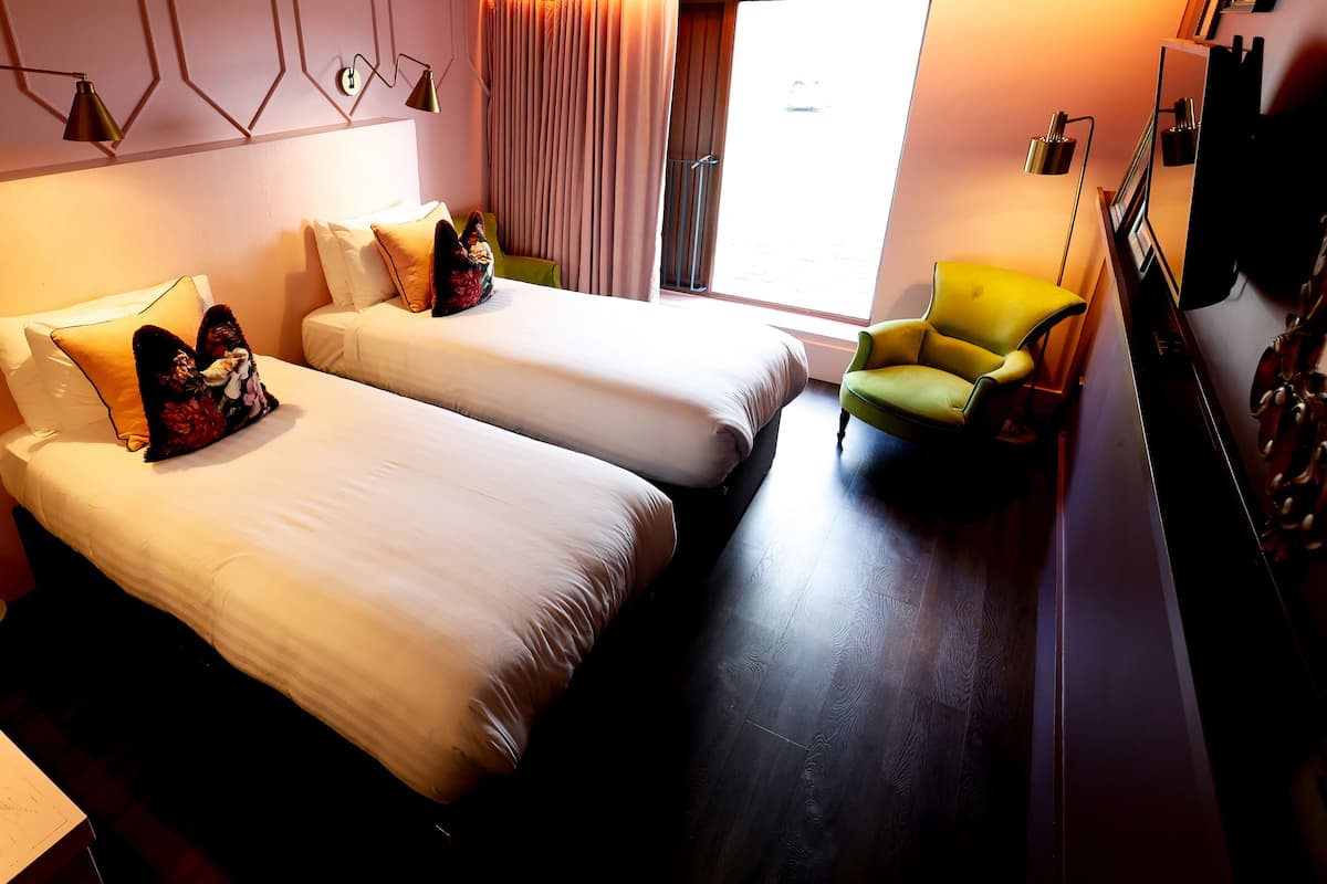 A hotel room with two beds, a green armchair, a large window with curtains, and a flat-screen TV mounted on the wall.