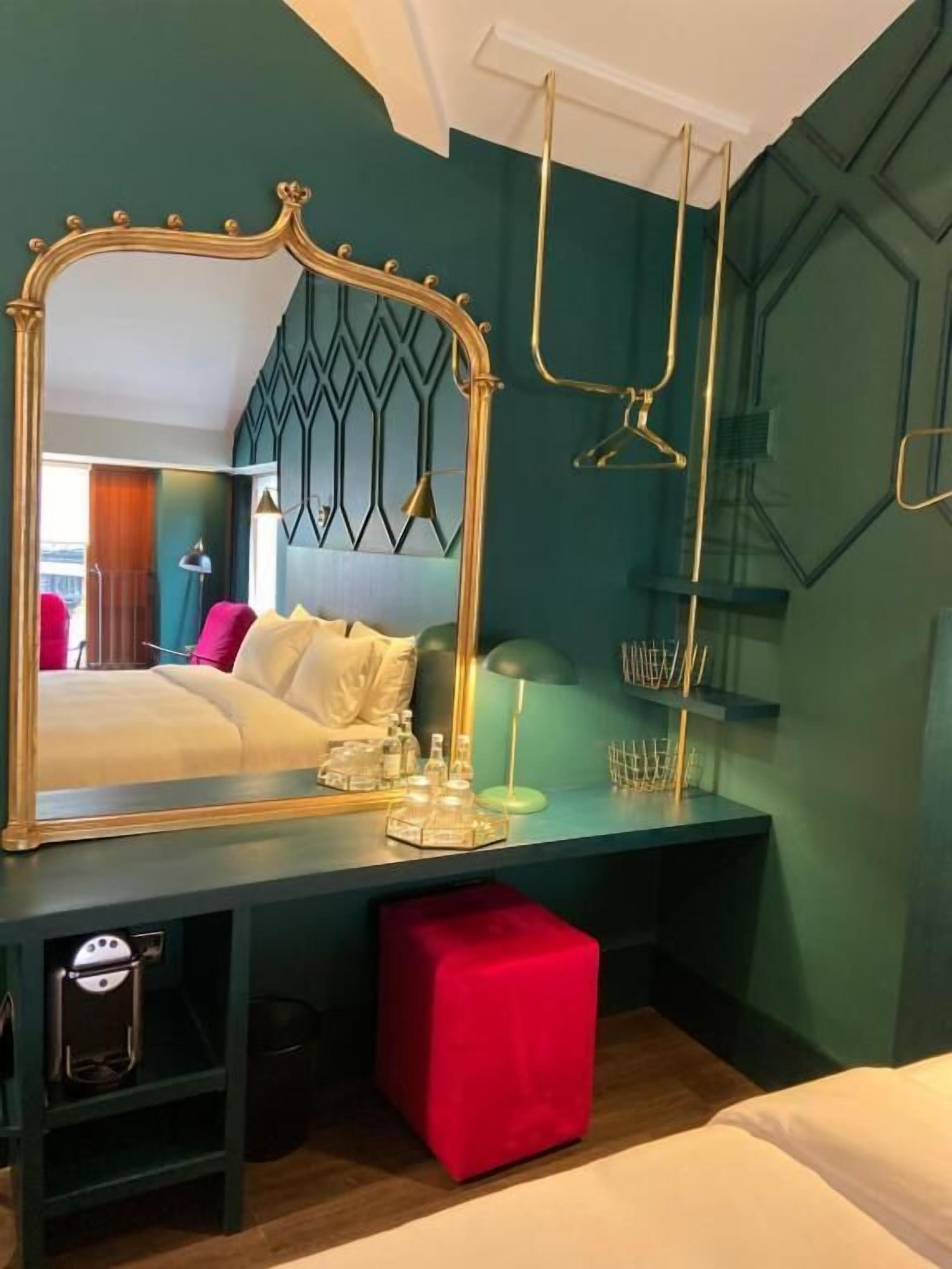 Habitación de hotel con un espejo grande, una cama con ropa blanca, un reposapiés rojo, un escritorio con lámpara y una lámpara colgante.