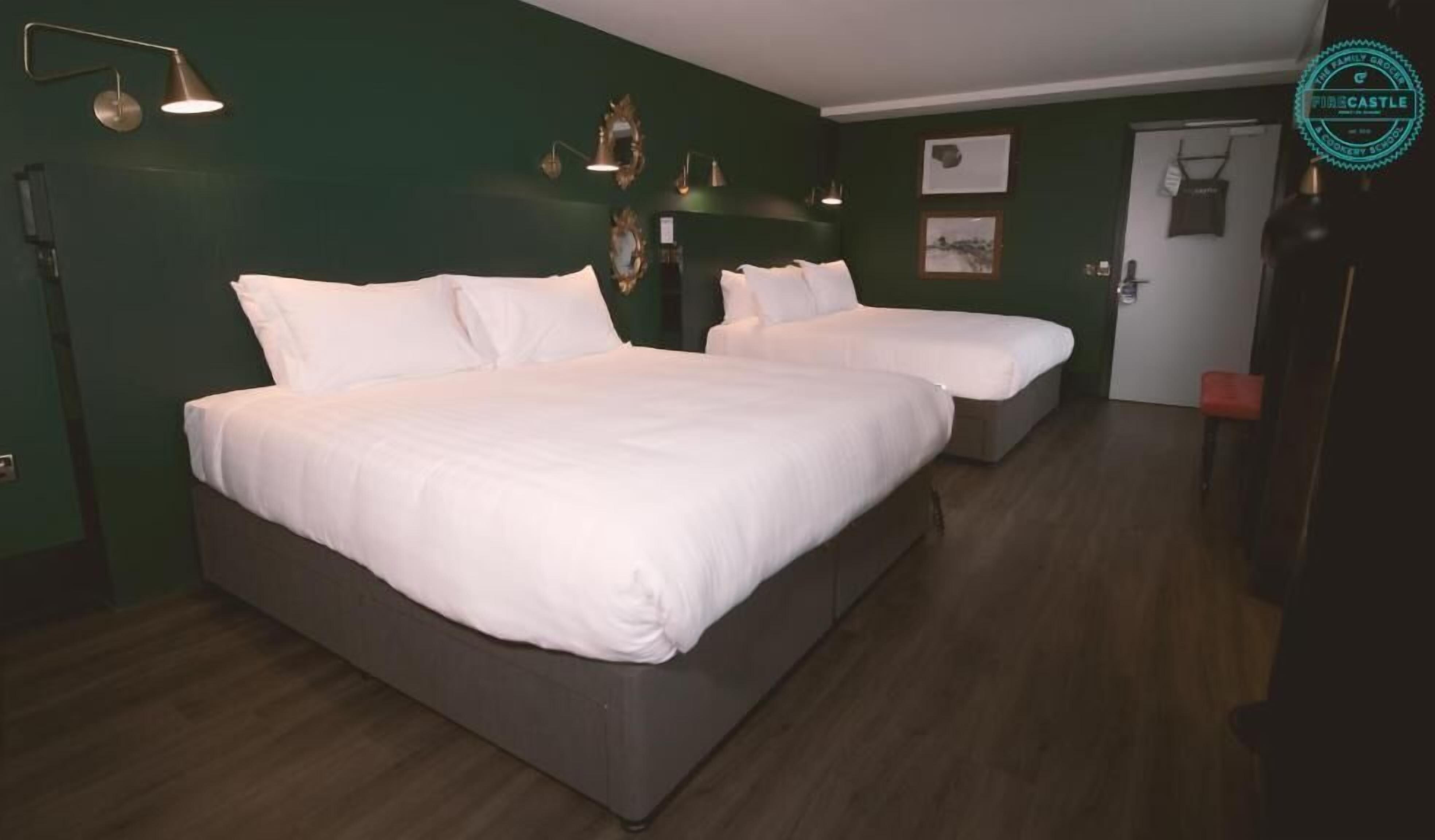 Habitación de hotel con dos camas, suelo de madera oscura, paredes verdes e iluminación instalada en la pared.
