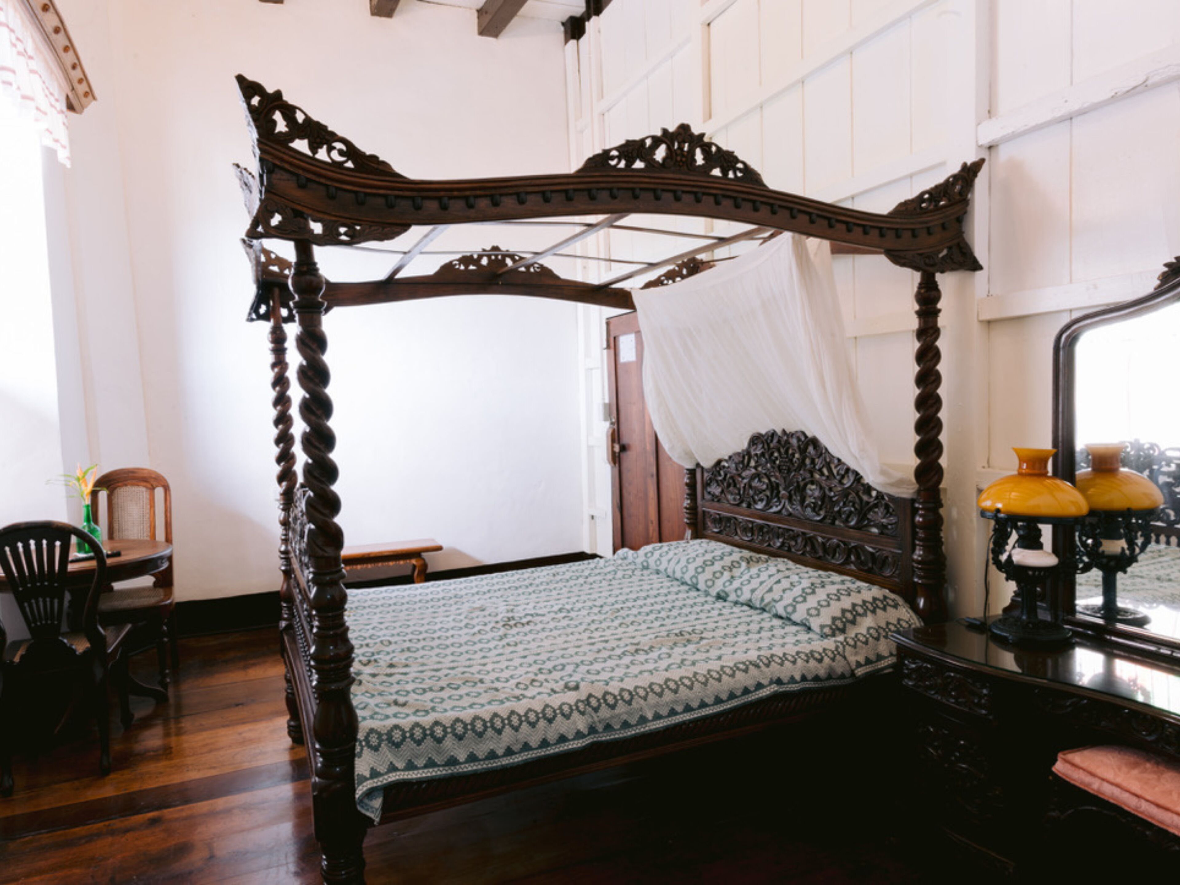Una cama con dosel con una colcha estampada, una silla de madera, una mesita y un espejo con lámparas amarillas.