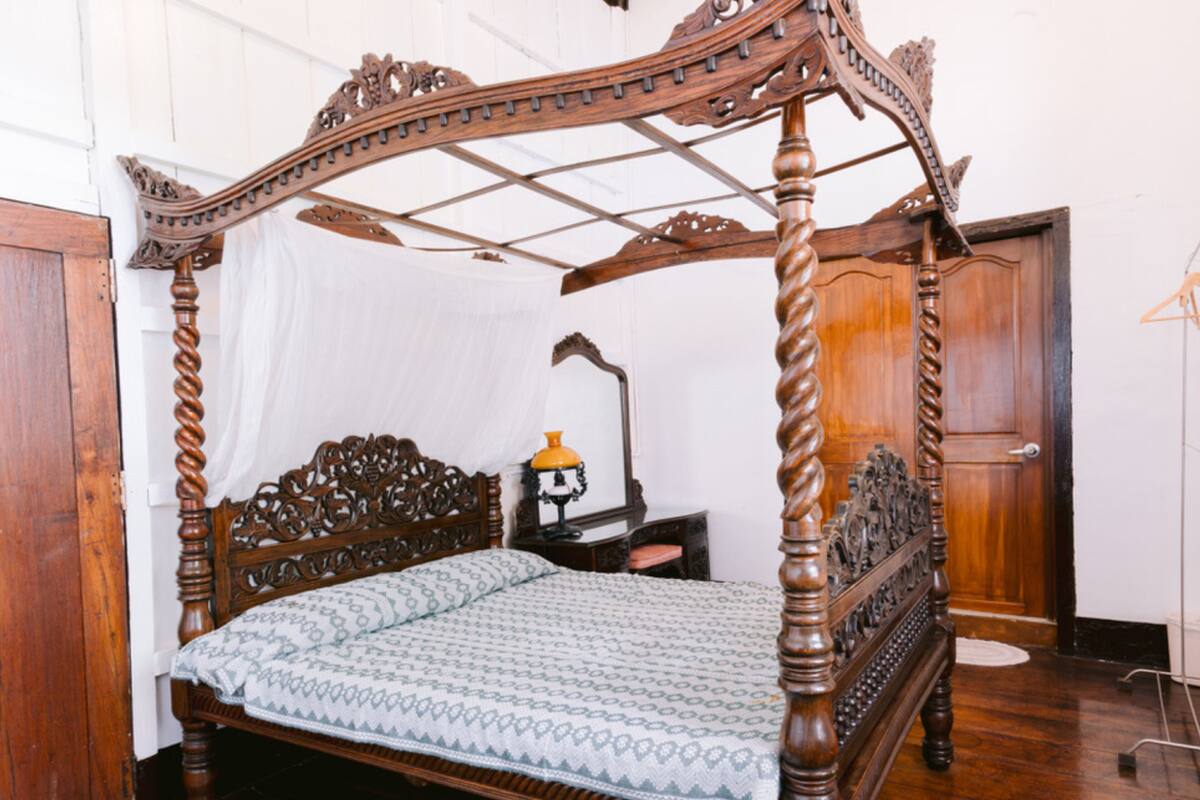 Una cama con dosel adornada con intrincados grabados, una mesita de noche con lámpara y un armario de madera en una habitación con paredes blancas y suelo de madera.