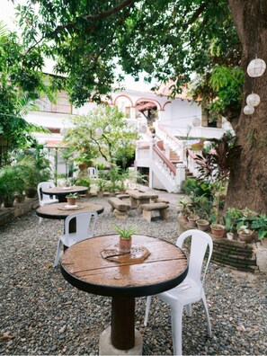 Ein Terrassenbereich im Freien mit runden Tischen und weißen Stühlen, umgeben von Kübelpflanzen und einem großen Baum.