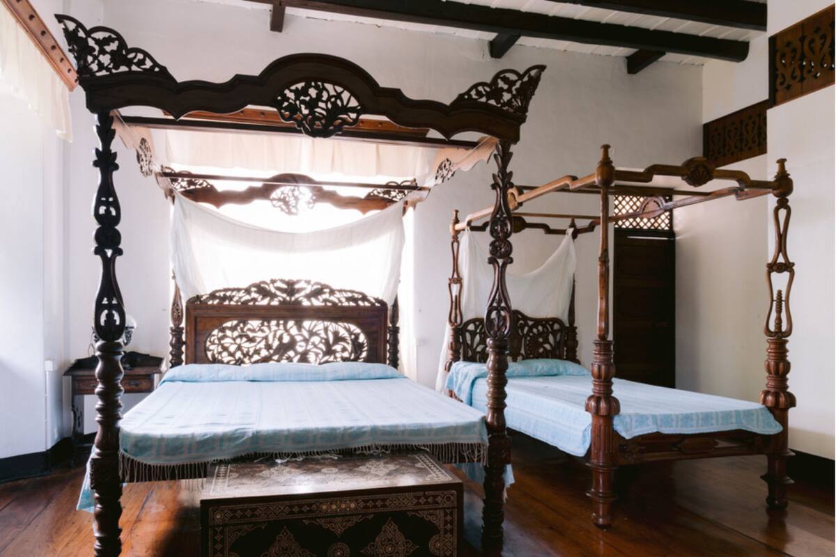 Una cama tradicional con dosel y dosel a juego, un sillón con dosel coordinado y una pequeña mesa decorativa en una habitación con suelo de madera y paredes blancas.