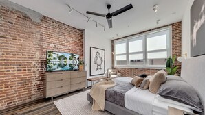 A bedroom with a brick wall, a large window, a bed with pillows, a desk, and a ceiling fan. - New Construction Loft Style Spacious Lux Apt 201 (Atlanta)
