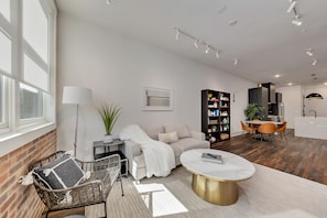 A modern living room with a sofa, a coffee table, and a wicker chair. There is a dining area with a table and chairs, a kitchen with a black cabinet, and a large window providing natural light.