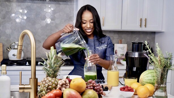 Una cocina con una persona sirviendo un líquido verde en un vaso, rodeada de frutas diversas y una máquina de jugos.