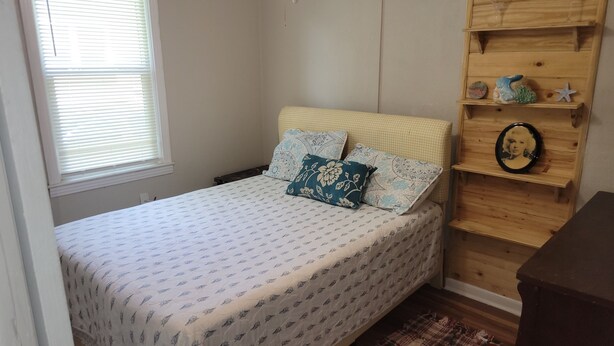 A bedroom with a bed, a window, a wooden shelf with decorative items, and a plaid rug. - Spencerville Center House (Tallahassee)