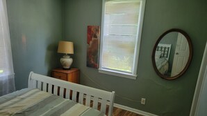 Un dormitorio con cama, mesita de noche, lámpara, ventana con persianas, un cuadro y un espejo.