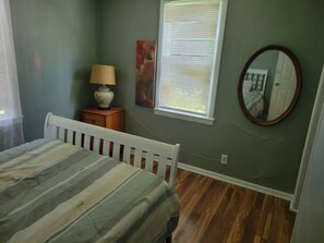 A bedroom with a bed, a nightstand, a lamp, a window with blinds, a painting, and a mirror. - Spencerville Center House (Tallahassee)