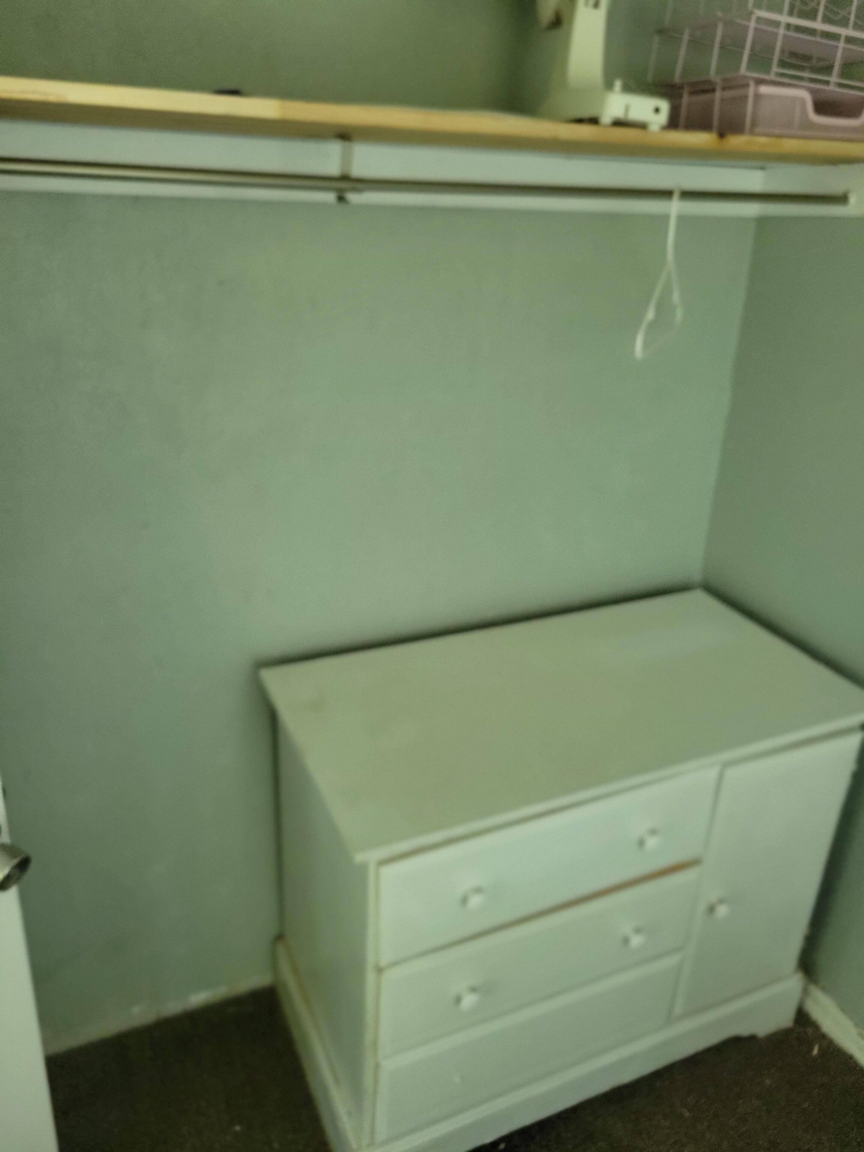A corner of a room with a white dresser, a shelf above it, and a wall with a light green color.