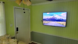 A room with a television mounted on the wall displaying a scenic landscape, a door, and a floral-patterned sofa. - Spencerville Center House (Tallahassee)