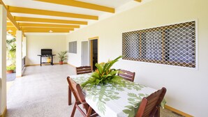 A dining area with a table set for four, surrounded by wooden chairs. There is a grill in the background, and the room has a yellow ceiling with white beams. - Muri Homeland Villa N2 (Ngatangiia District)