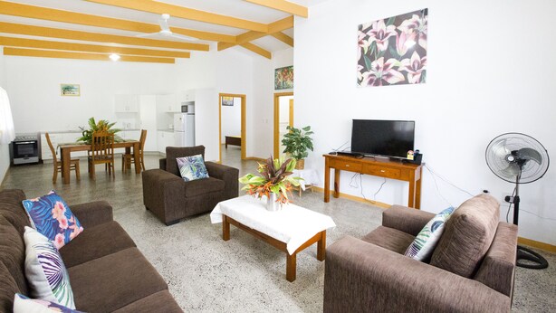 A living room with brown sofas, a coffee table, a TV, a fan, and a dining area with a table and chairs. - Muri Homeland Villa N2 (Ngatangiia District)