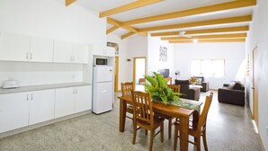 A compact kitchen with white cabinets and a white refrigerator, a wooden dining table with chairs, and a living area with a sofa and a television. - Muri Homeland Villa N2 (Ngatangiia District)