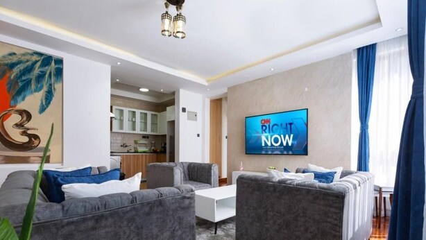 Ein modernes Wohnzimmer mit grauen Sofas, einem Flachbildfernseher und einer Küche im Hintergrund.