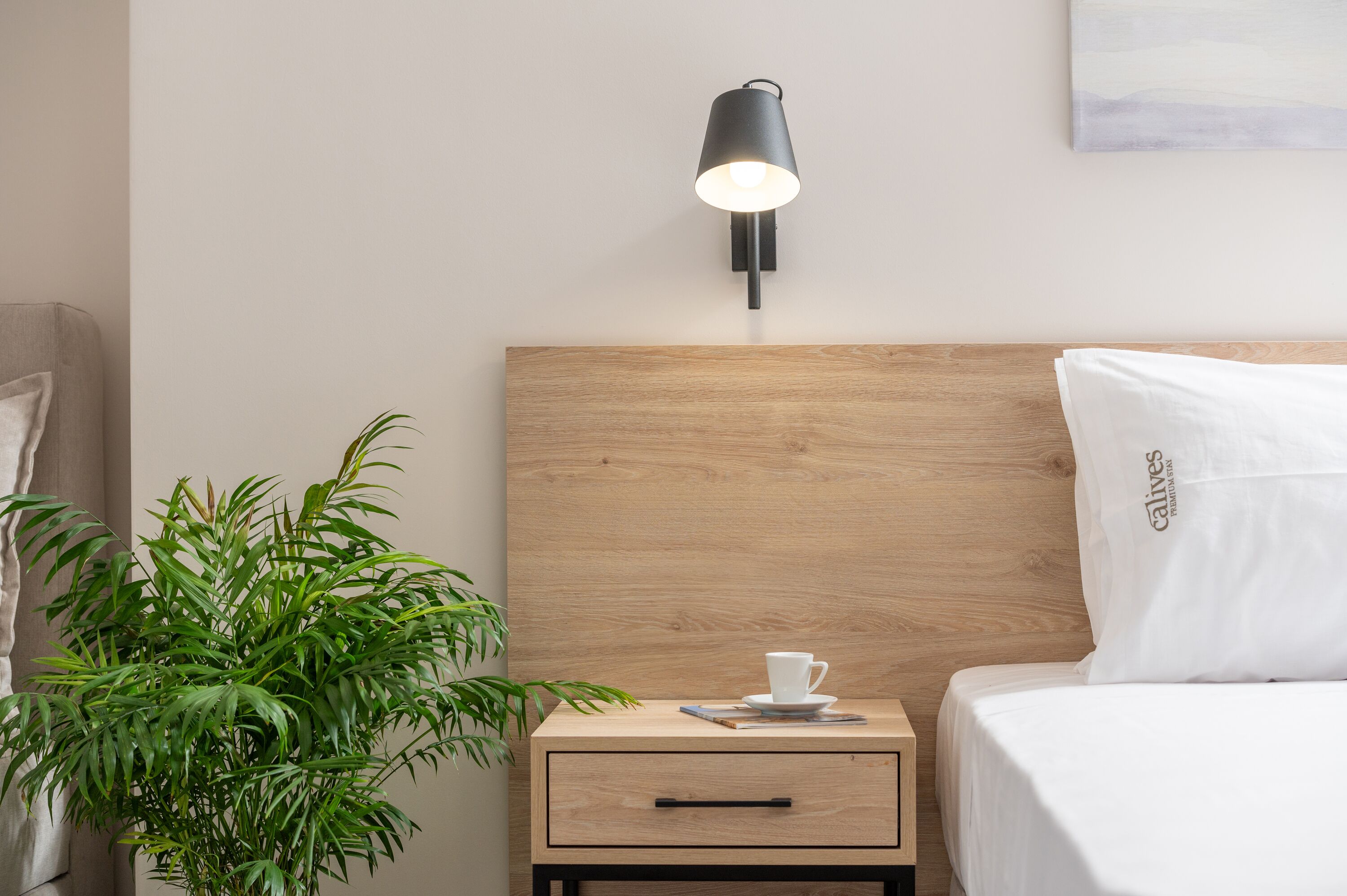 A neatly made bed with a white pillow, a bedside table with a cup and saucer, a small plant, and a wall-mounted lamp.
