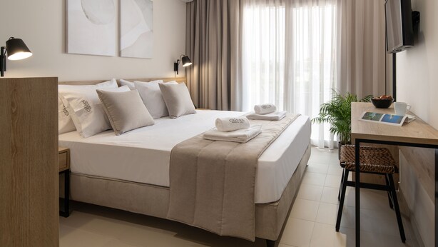 A hotel room with a bed, bedside lamps, a desk with a chair, a television, and a window with curtains. - Calives Premium Stay (Polygyros)