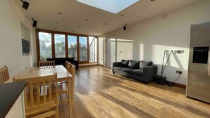 A spacious living area with a wooden dining table, a sofa, and a large window providing natural light. - Luxurious Two Bedroom Penthouse (London)