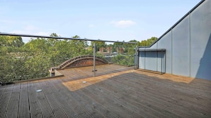 A rooftop terrace with a wooden deck, a glass balustrade, and a view of trees and buildings in the distance. - Luxurious Two Bedroom Penthouse (London)