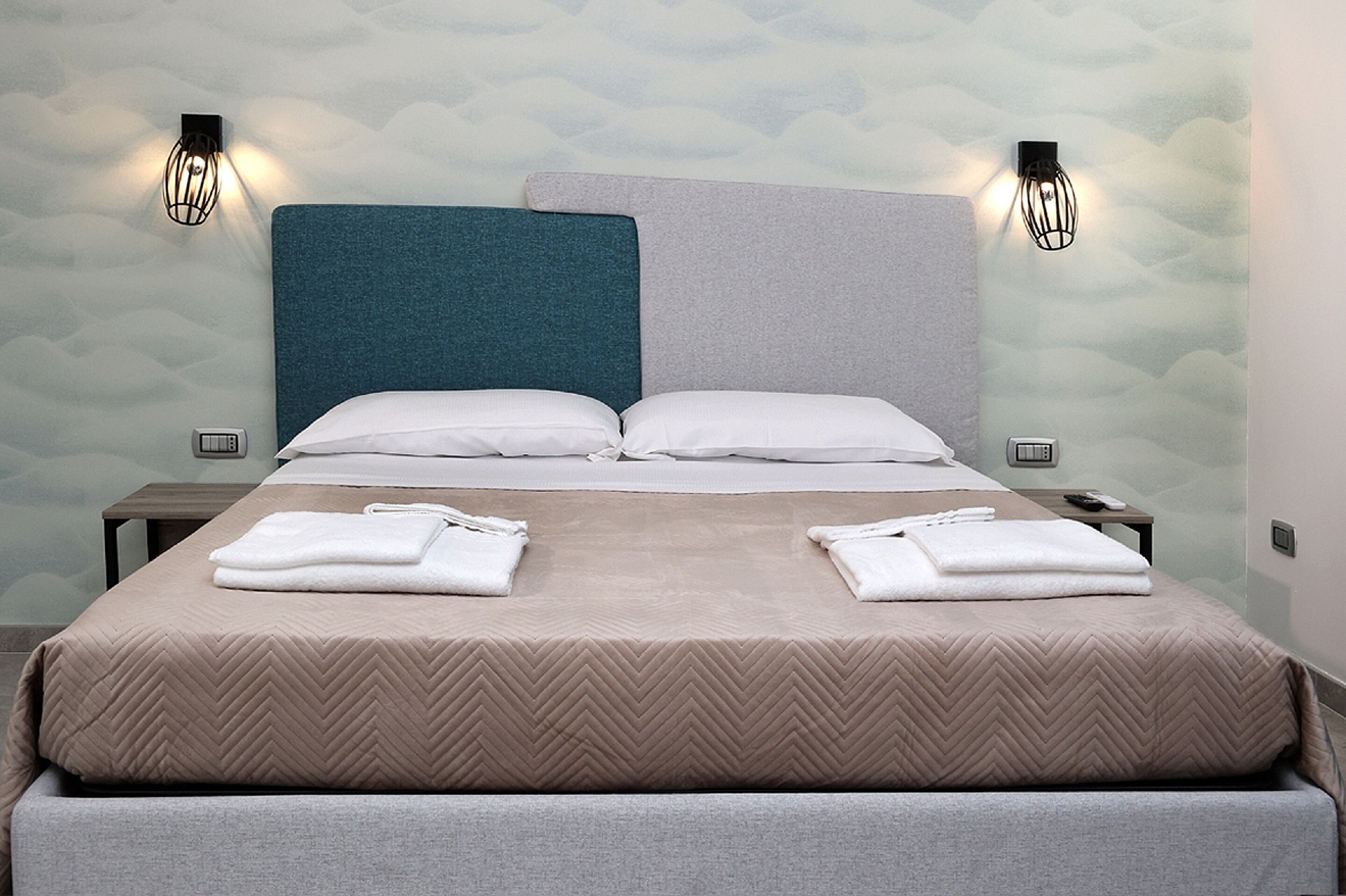 A neatly made bed with a herringbone patterned bedspread, two pillows, and folded towels on the side.