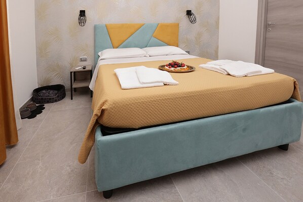Ein ordentlich gemachtes Bett mit gelb-blauer Kopfteil, ein kleiner Nachttisch mit Fernbedienung und ein Haustierbett in der Ecke.