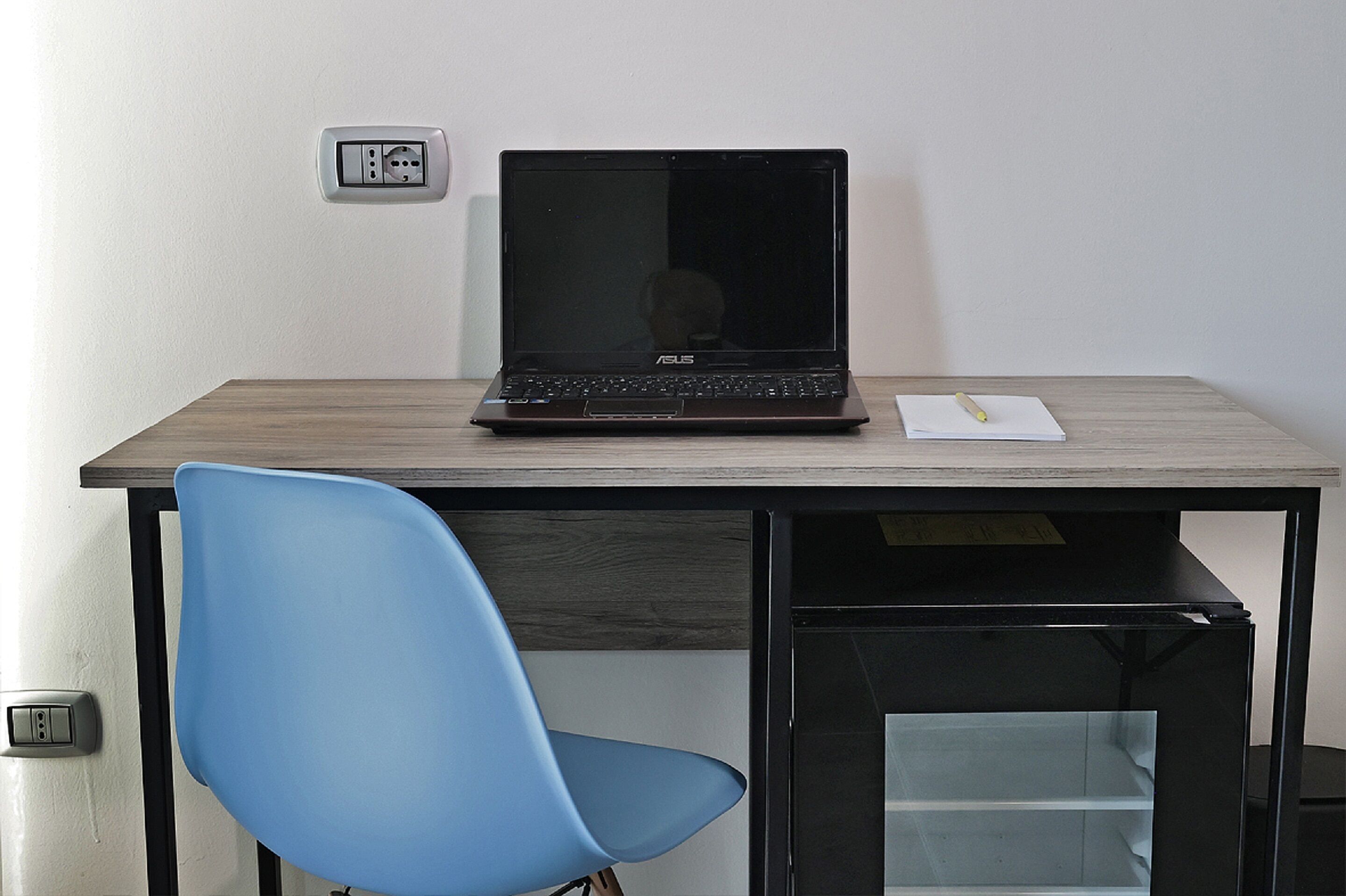 A laptop on a desk with a chair, a power outlet, and a trash bin.