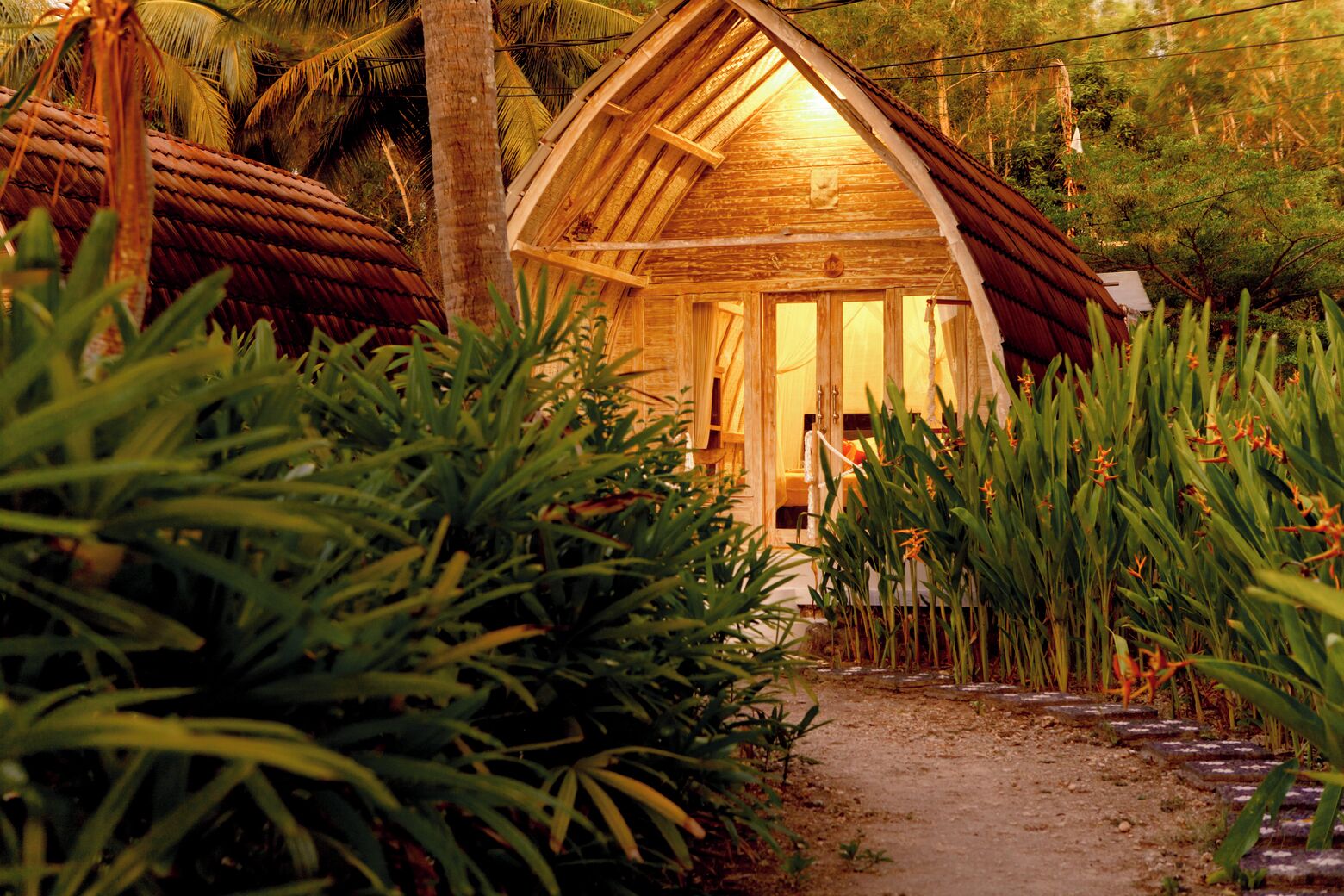 A rustic wooden cabin with a thatched roof, surrounded by lush greenery and a dirt path leading to the entrance.