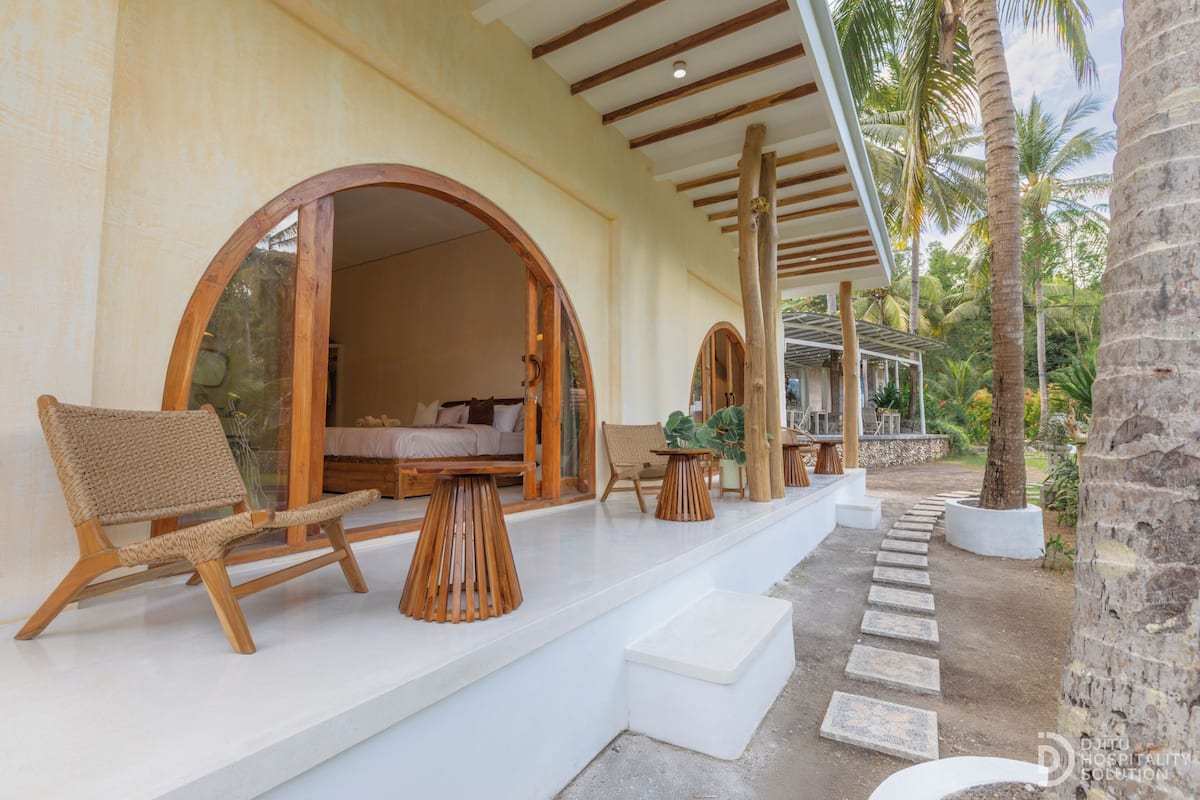 A covered outdoor seating area with wooden furniture and a stone pathway leading to a building entrance.