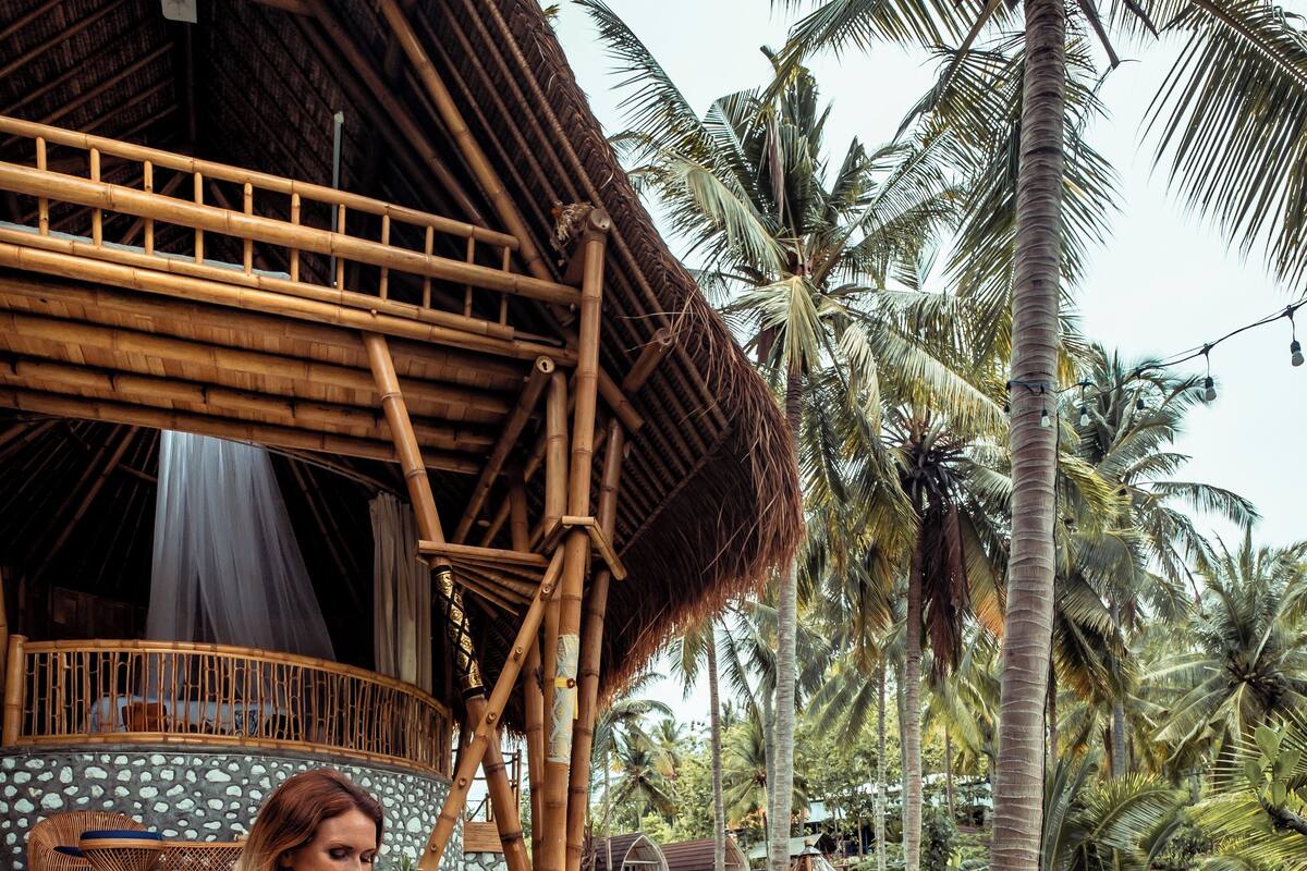 A woman sitting on a stone edge by a pool with a bamboo structure and palm trees in the background.