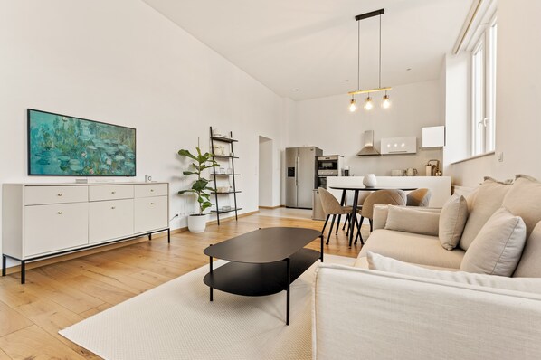 A modern living room with a sofa, a coffee table, and a TV. There is a kitchen area with a dining table and chairs.