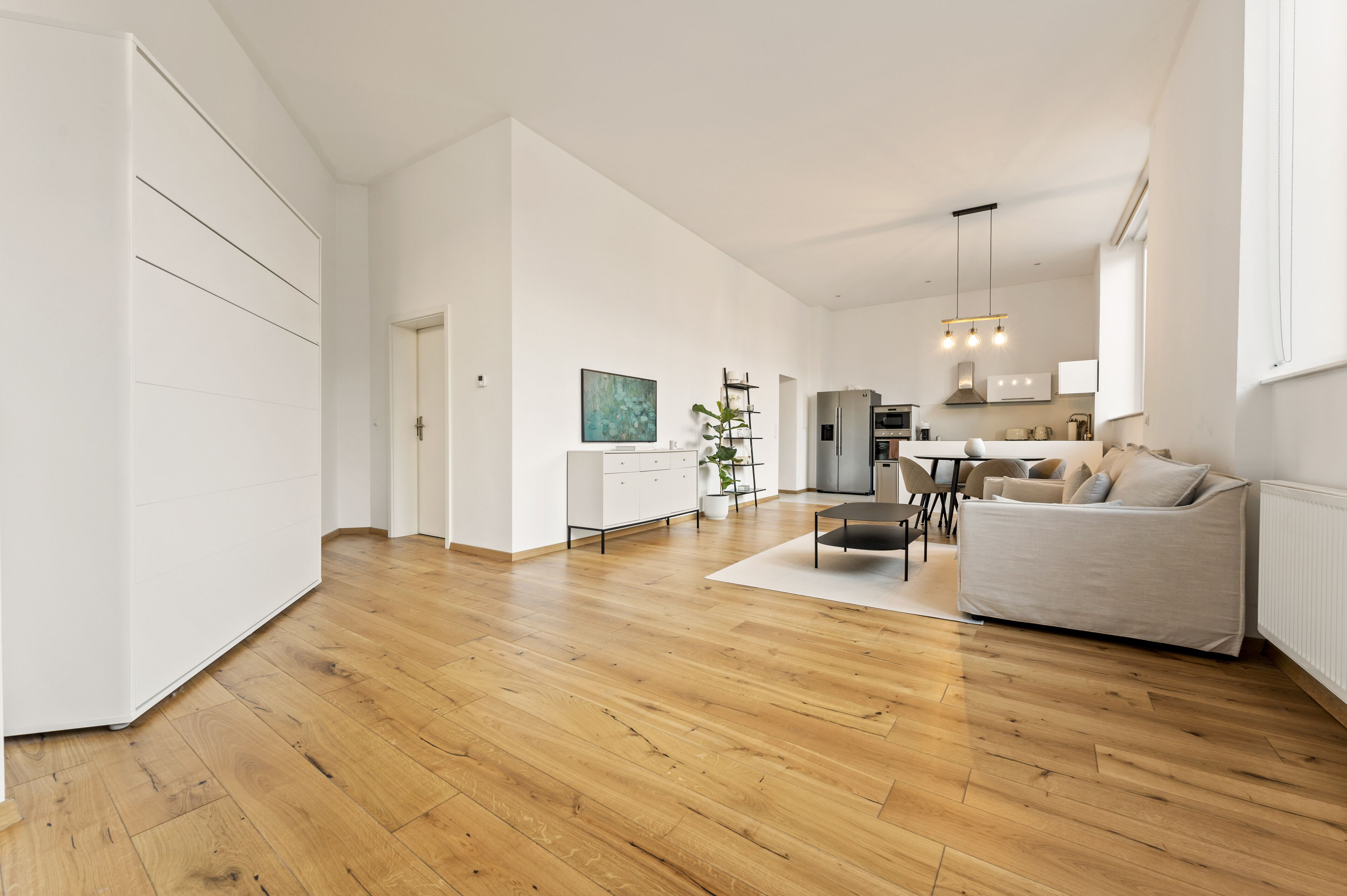 A spacious living area with a wooden floor, a white sofa, a dining table with chairs, and a kitchen in the background.