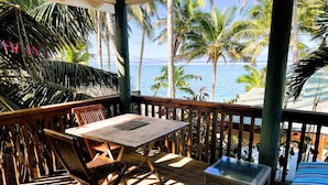 Eine Holzterrasse mit Tisch und Stühlen, mit Blick auf einen Strand mit Palmen und dem Meer.