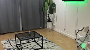 Un salon avec une table basse en verre, une plante en pot, un téléviseur à écran plat et un tapis à motifs.