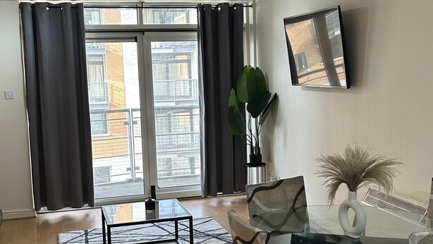 Un salon moderne avec une table à manger en verre, des canapés à motifs, une grande fenêtre avec des rideaux, un ventilateur de plafond et un téléviseur fixé au mur.