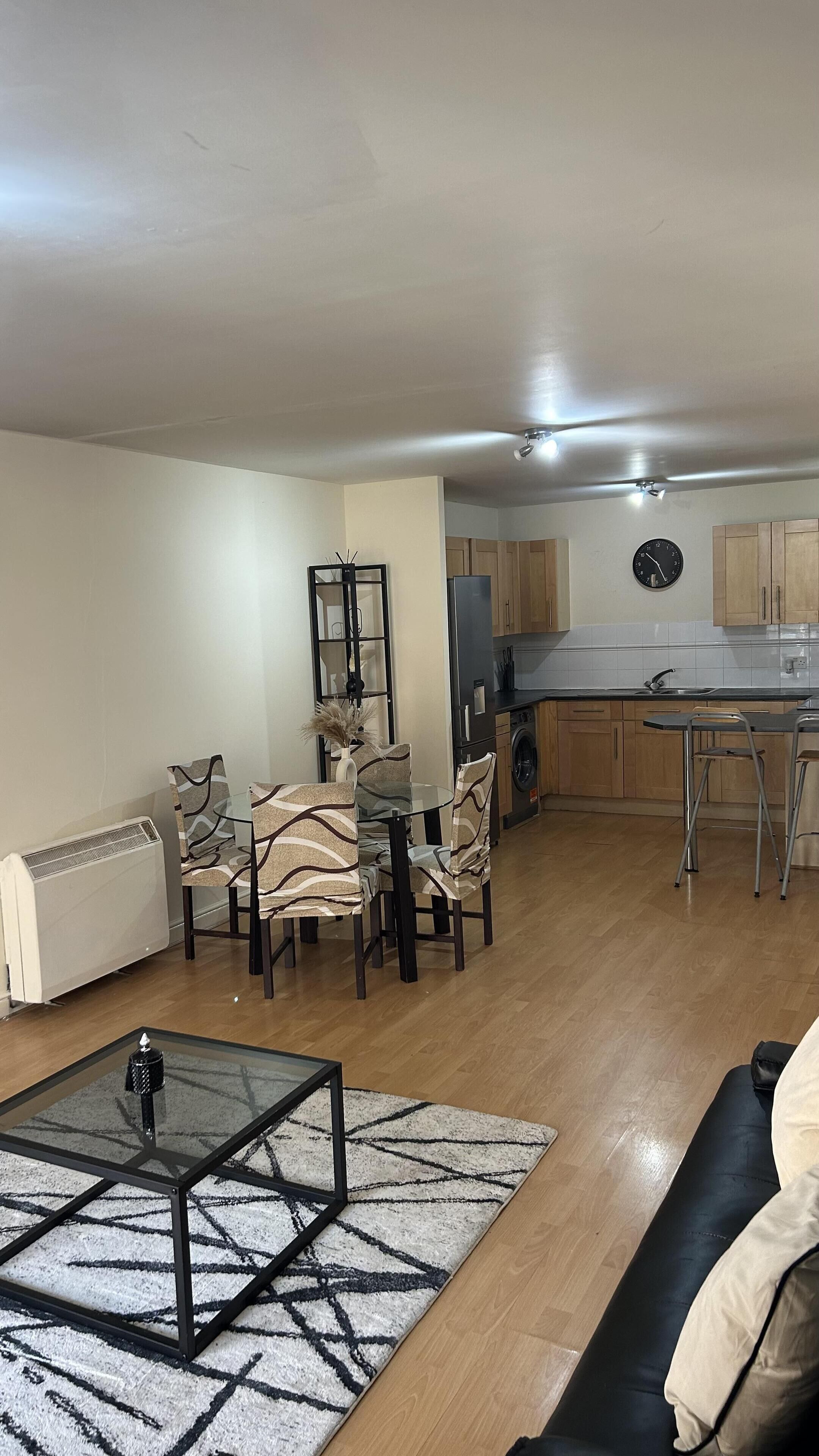 A living room with a glass coffee table, a sofa, patterned chairs, and a kitchen area with wooden cabinets and a sink.