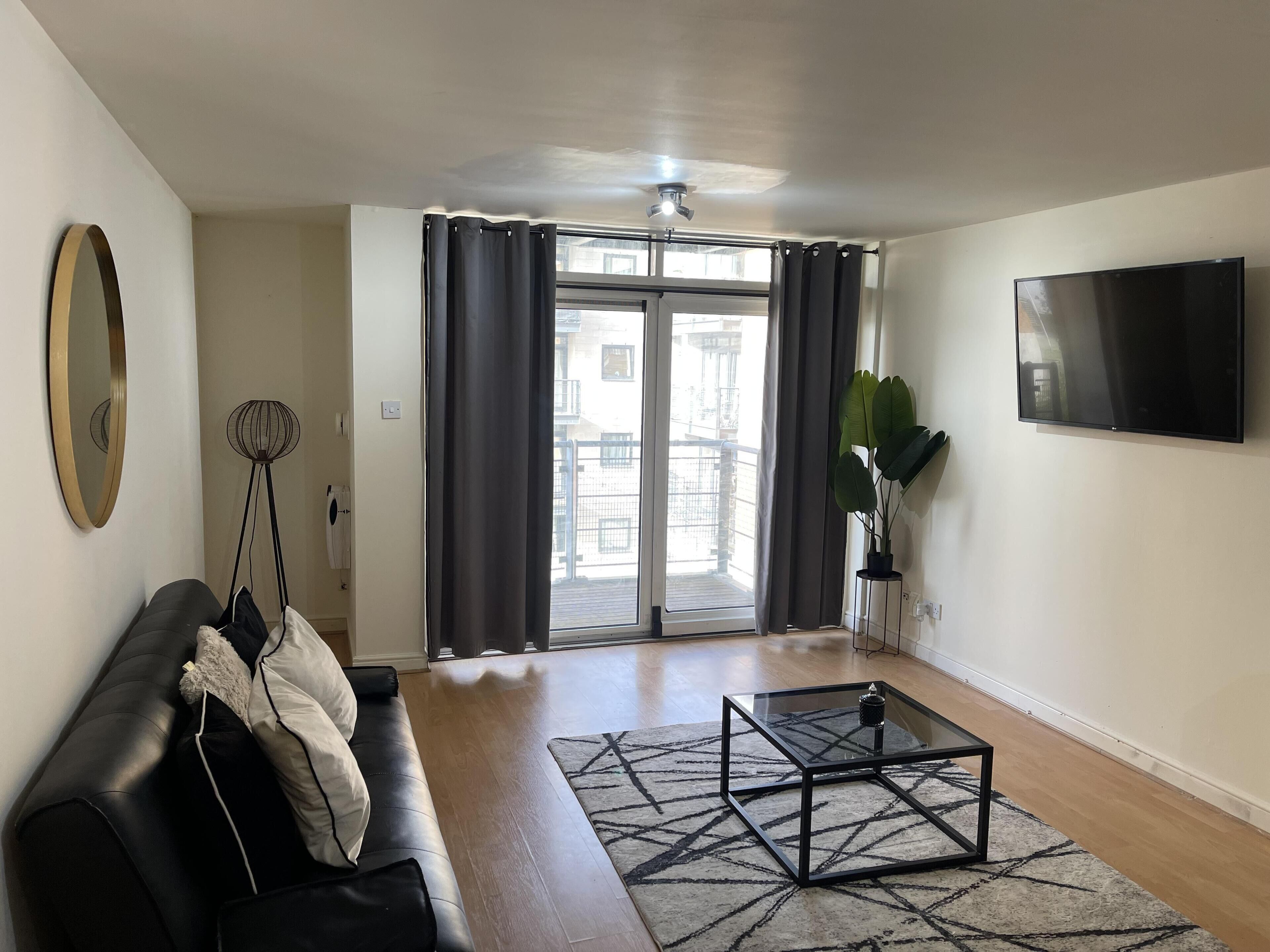 A modern living room with a black leather sofa, a glass coffee table, a large flat-screen TV, and a view of a balcony through a sliding door.