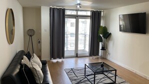 Un salon moderne avec un canapé en cuir noir, une table basse en verre, un grand téléviseur à écran plat et une vue sur un balcon accessible par une porte coulissante.