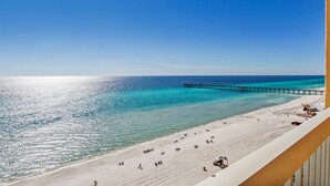 A beach with a pier extending into the ocean, clear skies, and a sandy shoreline with people and vehicles.