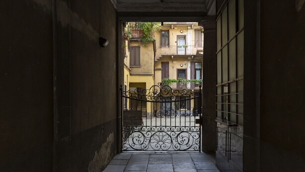 A narrow alleyway leading to a gated courtyard with buildings in the background. - nota-Mi - Domus Ticinese (Milan)